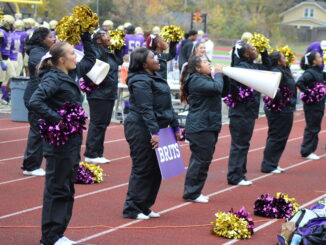 A group of girls varying in race and height stand in two horizontal lines on the sidelines of a football field to cheer. They’re clad in black jumpsuits, holding metallic purple and gold pompoms. One of the girls in front holds a megaphone to her mouth, while the girl beside her has a purple sign with the word ‘BRITS’ written in white.