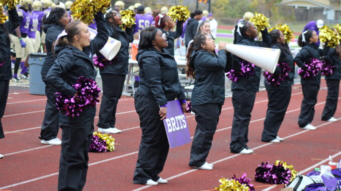 A group of girls varying in race and height stand in two horizontal lines on the sidelines of a football field to cheer. They’re clad in black jumpsuits, holding metallic purple and gold pompoms. One of the girls in front holds a megaphone to her mouth, while the girl beside her has a purple sign with the word ‘BRITS’ written in white.