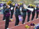 A group of girls varying in race and height stand in two horizontal lines on the sidelines of a football field to cheer. They’re clad in black jumpsuits, holding metallic purple and gold pompoms. One of the girls in front holds a megaphone to her mouth, while the girl beside her has a purple sign with the word ‘BRITS’ written in white.