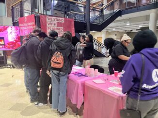 A group of college students stand in front of a pink table holding plastic cups, straws and napkins. Behind the table, other students watch them examine the items on the table.