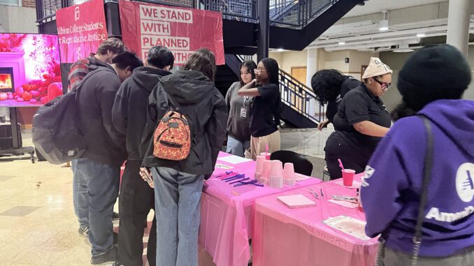 A group of college students stand in front of a pink table holding plastic cups, straws and napkins. Behind the table, other students watch them examine the items on the table.
