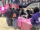 A group of college students stand in front of a pink table holding plastic cups, straws and napkins. Behind the table, other students watch them examine the items on the table.