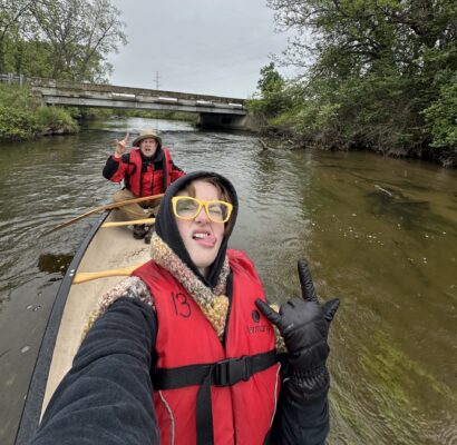A selfie of two people in red life-jackets sitting on a canoe on a river. The person in front, holding the camera, sticks out their tongue, and both hold up a rock-and-roll gesture with one hand.