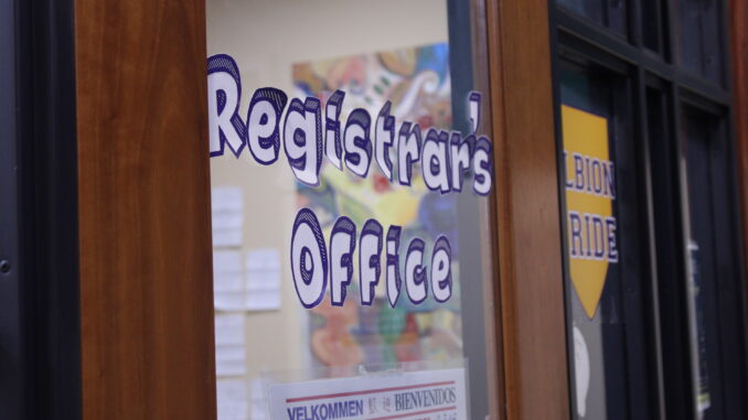 Paper letters reading “Registrar’s Office” are stuck to the glass of a wood-rimmed door. Through the glass, a room with papers and a painting on the wall is visible.