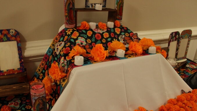A close up shot of a rectangular table covered by both white and black table clothes. There are multiple layers of the display on the table; on it are picture frames, fake candles, real candles, and strings of balls of orange paper.