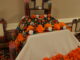 A close up shot of a rectangular table covered by both white and black table clothes. There are multiple layers of the display on the table; on it are picture frames, fake candles, real candles, and strings of balls of orange paper.