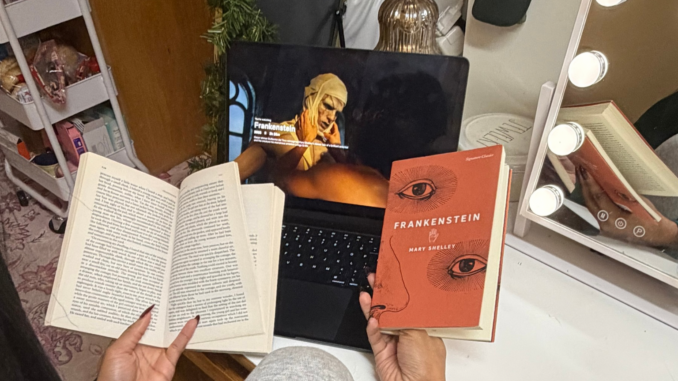 An individual sits at a desk, with their head turned away from the camera. The individual is holding out both hands, both holding one book each. The book on the right is closed and the title “Frankenstein” is visible, while the book on the left is open. Behind both books is a laptop open to Netflix, paused on a movie scene.