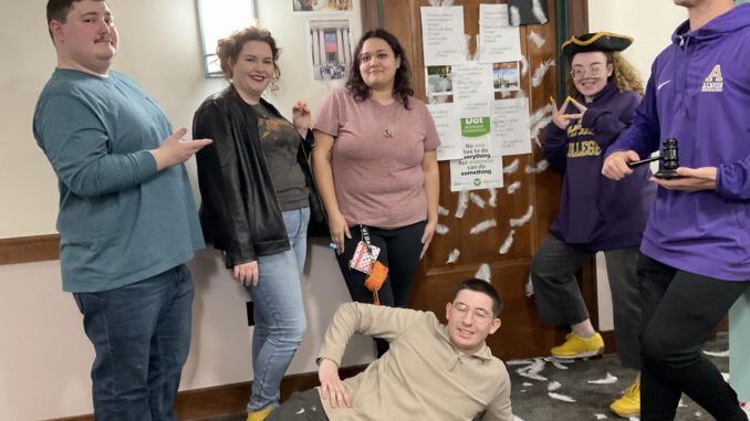 A group of six people posing in a hallway with a decorated door behind them. The person in the middle is laying on the floor with one hand propped on a hip.