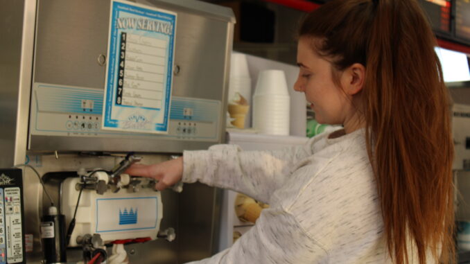 A girl with a blonde ponytail and a nose piercing holds down the lever on an ice cream machine to make a swirl of chocolate and vanilla into a cone.