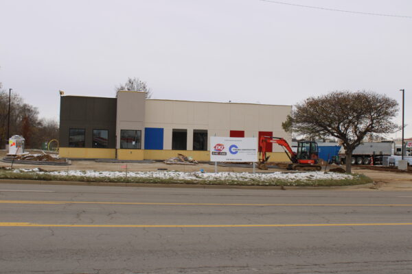 A tan building with red and blue paneling. In front of the building is an orange crane and a sign in front of it that reads “another quality project by Com-Res Construction.”
