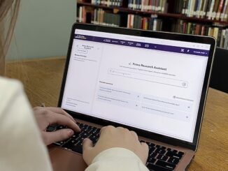 An open laptop sits on a wooden table; the computer screen reads “Primo Research Assistant.” A woman’s hands are on the keyboard, and there are shelves of books in the background.