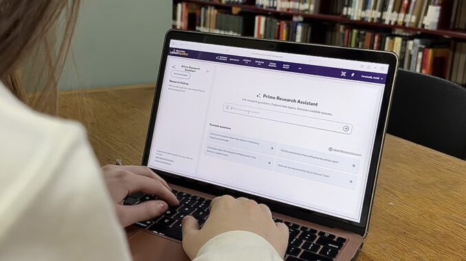 An open laptop sits on a wooden table; the computer screen reads “Primo Research Assistant.” A woman’s hands are on the keyboard, and there are shelves of books in the background.
