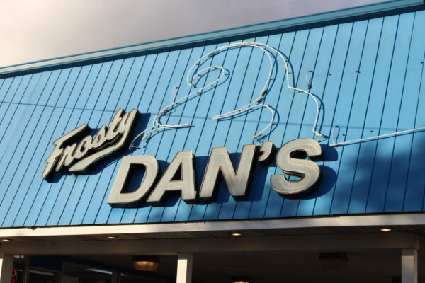 A sign set against the siding of a blue roof reads “Frosty Dan’s,” with the outline of a swirl of ice cream on top of the word “Dan’s” in unlit neon lights.