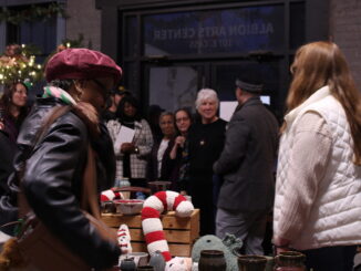 A group of people gather in a gray brick-walled room filled with varying art pieces and surrounded with festive decor for the holidays.