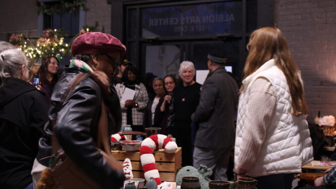 A group of people gather in a gray brick-walled room filled with varying art pieces and surrounded with festive decor for the holidays.