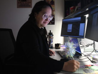 An individual with black pigtails sits in front of a desk, head tilted down toward it. The person is holding a sparkly white pen and writing on a notepad. In front of them are two computer monitors and a laptop, all with screens on.