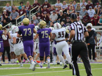 A man wearing a striped black and white shirt in the foreground, out of focus, runs towards a crowd of football players running in the same direction away from the camera. People sitting on bleachers are visible in the background.