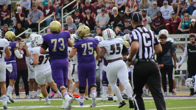 A man wearing a striped black and white shirt in the foreground, out of focus, runs towards a crowd of football players running in the same direction away from the camera. People sitting on bleachers are visible in the background.