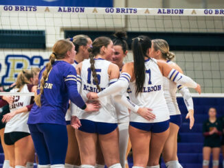 A huddle of volleyball players congregate together mid-match. In the background, the top of a volleyball net and several students sitting in bleachers can be seen.