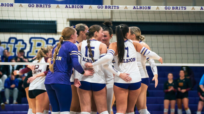 A huddle of volleyball players congregate together mid-match. In the background, the top of a volleyball net and several students sitting in bleachers can be seen.