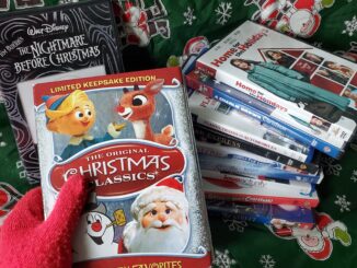 A hand with a red fuzzy glove holds an ornate box with white text reading “The Original Christmas Classics.” Behind the box is a large, leaning stack of several DVD cases to the right and a copy of “The Nightmare Before Christmas” to the left leaning against a green backdrop with snowflakes and cartoon Santa Clauses printed on it.