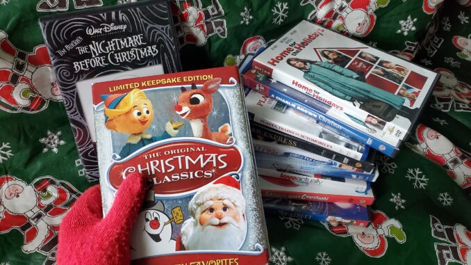 A hand with a red fuzzy glove holds an ornate box with white text reading “The Original Christmas Classics.” Behind the box is a large, leaning stack of several DVD cases to the right and a copy of “The Nightmare Before Christmas” to the left leaning against a green backdrop with snowflakes and cartoon Santa Clauses printed on it.