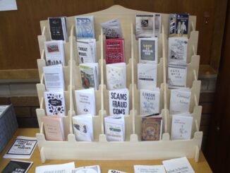 A tiered grid of small wooden shelves hold folded paper books. On the table the display stands on, other small paper magazines are scattered around.