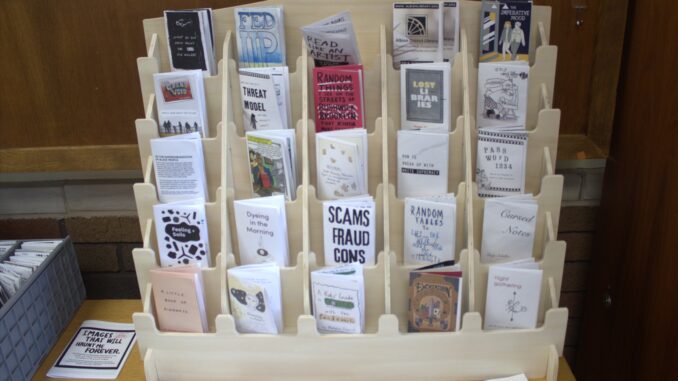 A tiered grid of small wooden shelves hold folded paper books. On the table the display stands on, other small paper magazines are scattered around.