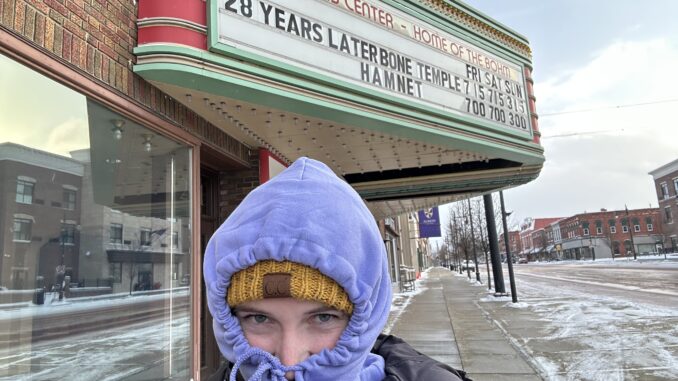 A woman in a black winter coat stands with a purple sweatshirt hood tied over a yellow hat. Behind her is a sign for a movie theatre that reads, “The Davis Center - Home of the Bohm,” and below are two movies and their showtimes including “28 Years Later: Bone Temple” and “Hamnet.” To the right of the sign is a downtown street with snow-covered sidewalks.