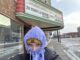 A woman in a black winter coat stands with a purple sweatshirt hood tied over a yellow hat. Behind her is a sign for a movie theatre that reads, “The Davis Center - Home of the Bohm,” and below are two movies and their showtimes including “28 Years Later: Bone Temple” and “Hamnet.” To the right of the sign is a downtown street with snow-covered sidewalks.