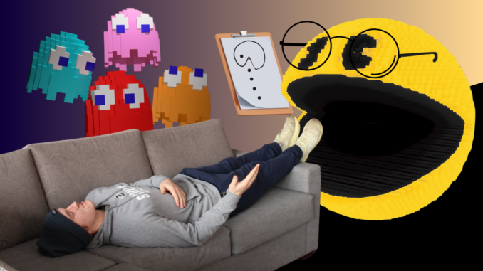A young man lies on a gray couch, feet up on the arm and gesturing as he speaks. Behind him are four pixelated multicolored ghosts, who look over him. At his feet is a 3D pixelated Pac-Man wearing glasses and a clipboard with a picture of Pac-Man drawn on it.