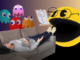 A young man lies on a gray couch, feet up on the arm and gesturing as he speaks. Behind him are four pixelated multicolored ghosts, who look over him. At his feet is a 3D pixelated Pac-Man wearing glasses and a clipboard with a picture of Pac-Man drawn on it.