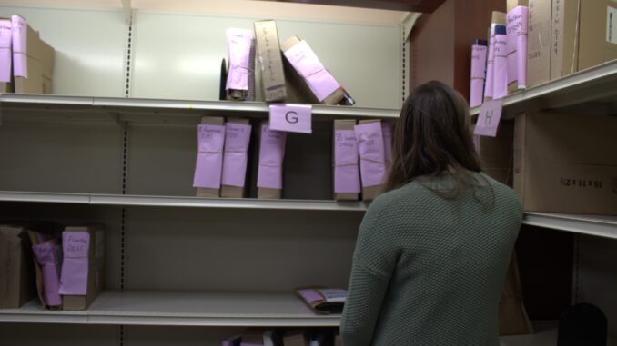 A student stands with their back to the camera, facing a wall of metal shelves. On the shelves are several brown cardboard boxes, stood up next to each other in alphabetical order. Purple slips of paper are attached to each box with a rubber band. A large purple label with the letter “G” written on it hangs from the shelf the student faces, and the letter “H” is written on a label on the shelf to their right.