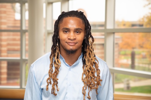 A professional headshot of a young man. The man is looking at the camera and is wearing a light blue dress shirt, with his hair down. The man additionally has a silver septum piercing and earrings.