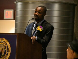 A young man dressed in a suit with a yellow ribbon and purple and yellow flowers as a boutonniere stands at a microphone, speaking. The microphone stand is inscribed with “Albion College” and a crest.