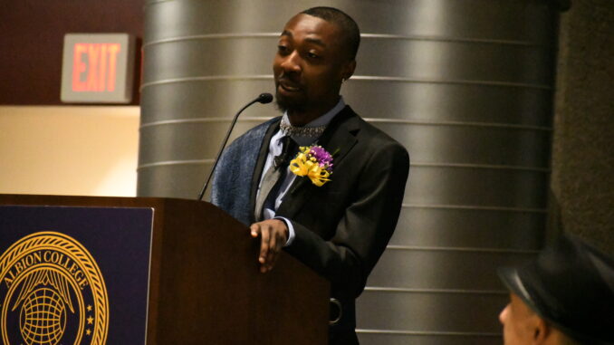A young man dressed in a suit with a yellow ribbon and purple and yellow flowers as a boutonniere stands at a microphone, speaking. The microphone stand is inscribed with “Albion College” and a crest.