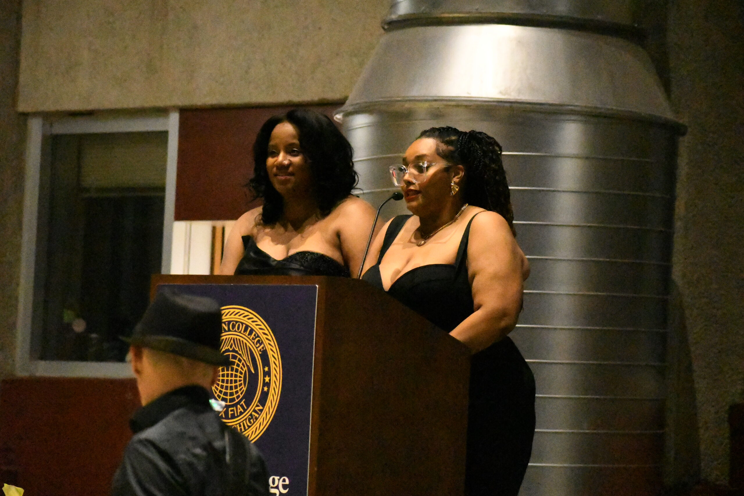 Two women in black dresses stand at a pedestal with a microphone. The woman on the right is speaking into the microphone. Beside them is a man in a red suit holding white roses.