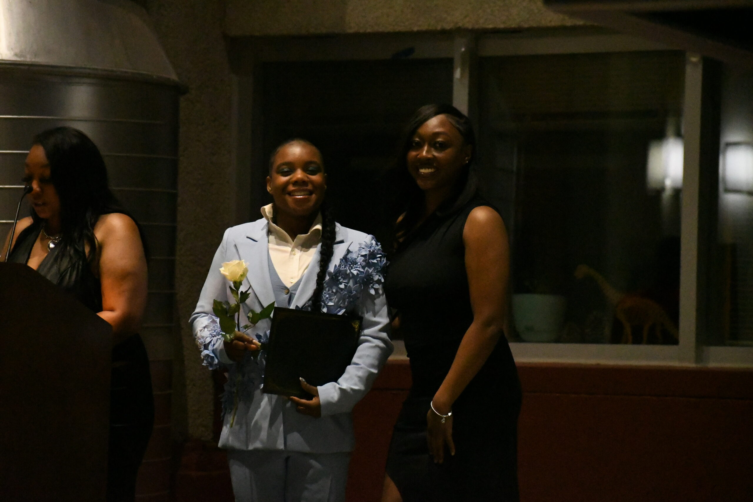 Two young women smile at the camera, with one in a light blue suit holding a white rose and an award. To her right is a woman in a black dress. The lighting is dim as they stand in front of a window.