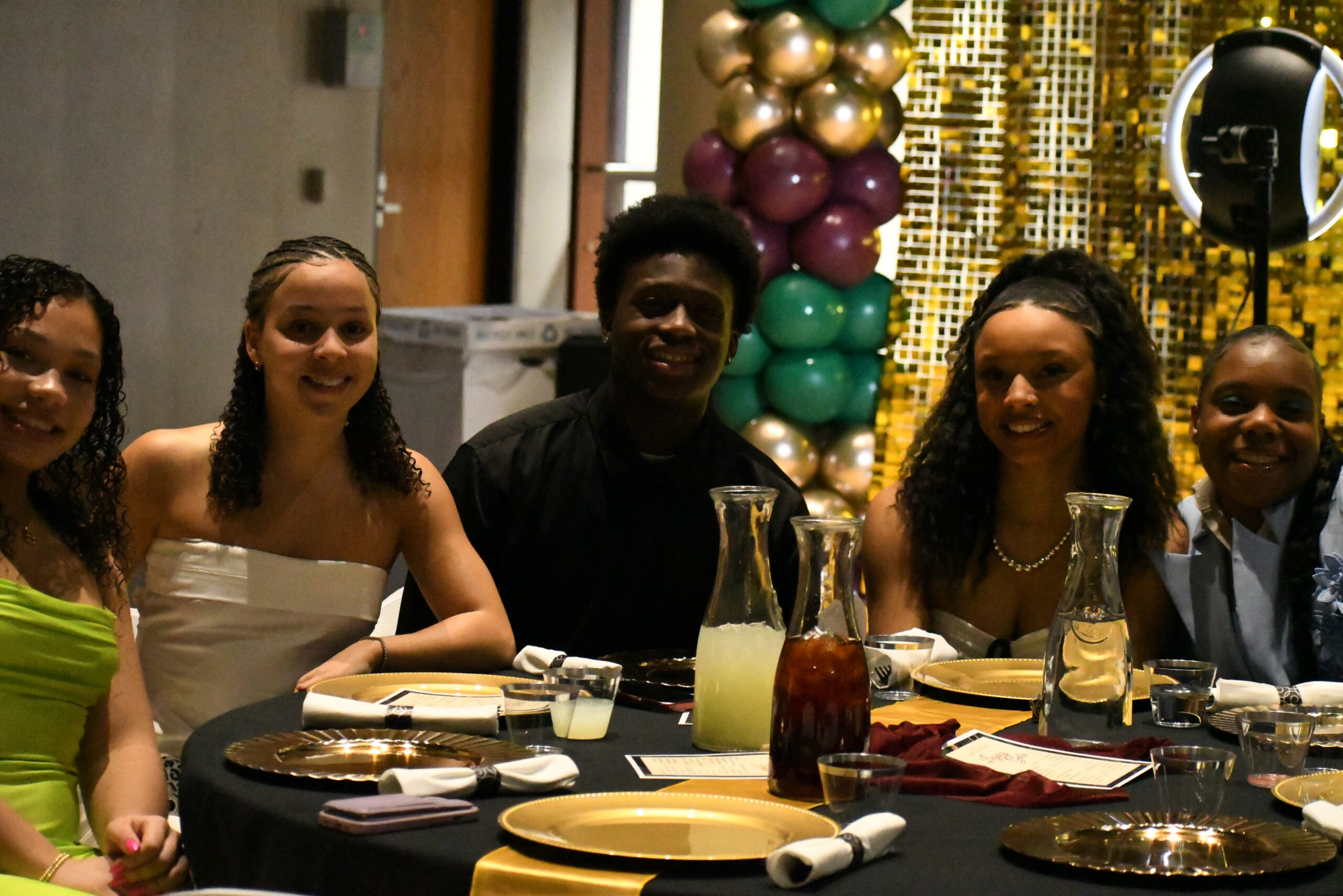 A group of five people sits around a table dressed for a formal event. A backdrop of colorful balloons and a reflective wall is shown behind them.