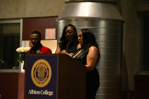 Two women in black dresses stand at a pedestal with a microphone. The woman on the right is speaking into the microphone. Beside them is a man in a red suit holding white roses.