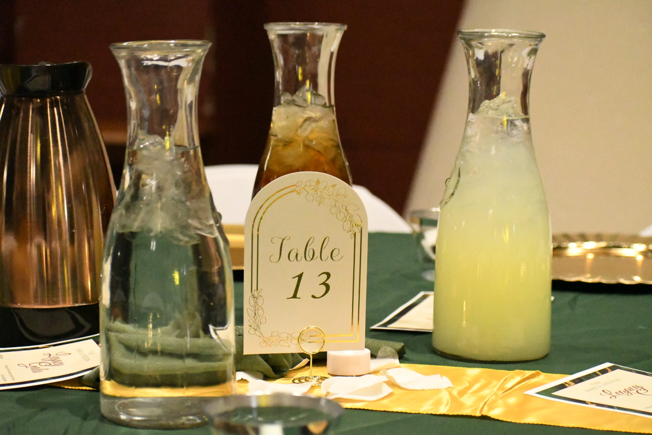 A round table is decorated with a dark green tablecloth with various gold details, such as pitchers and plates. On top of the table is a card labeled “Table 3.” Behind the card are 3 glass bottles, each containing water with ice, iced tea, or lemonade. Around the bottles are white rose petals and event pamphlets.