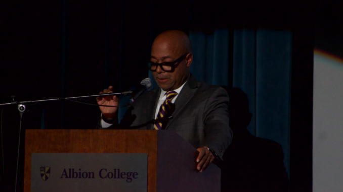 An older Black man stands behind a wooden podium with a silver plate reading “Albion College” and the school logo. He is wearing a gray suit with a white button-up shirt and a purple-and-gold tie with a blue curtain behind him.