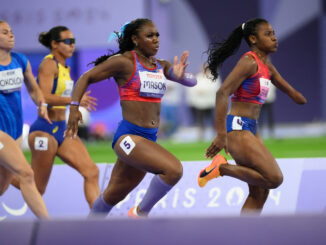 Four women sprint down an indoor track. The two women in front are wearing red and blue track singlets and shorts, with race bibs pinned to their uniforms. The other two women, running slightly behind, are also wearing track uniforms and racing bibs.