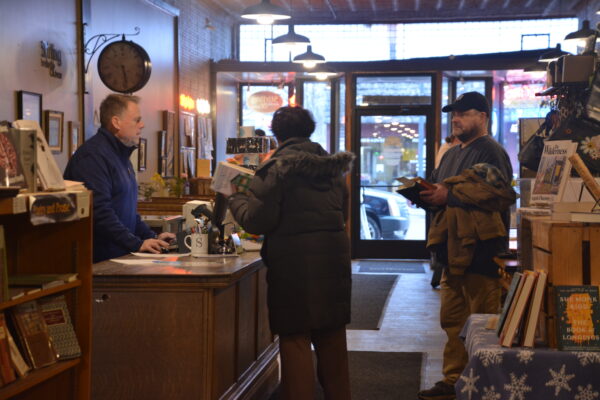 A medium-length shot of a man behind a desk at a cash register, checking out a woman in a black coat. To the right, a man stands in line behind the woman.