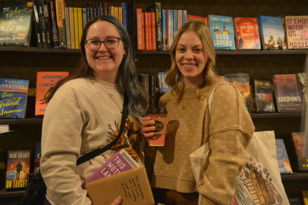 Two women pose, smiling, for a picture. The one on the right is wearing a yellow sweater with butterflies on it, holding a cup. The woman on the left is holding two books. There is a wall of bookshelves behind them. 