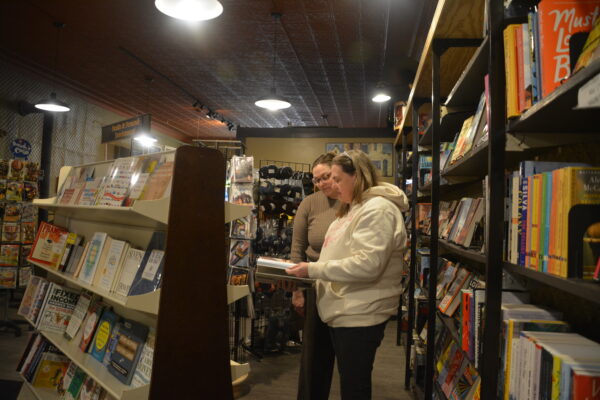 Two women look at a book together. In front of them is a bookcase at an angle, and behind them there is a long bookcase along the wall. Skeins of yarn hang on a rack in the background. 