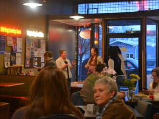 Many people stand and sit in an open, low-lit cafe. Parked cars are visible through the front windows of the store at the front of the room.