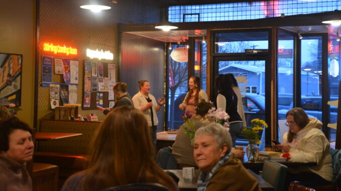 Many people stand and sit in an open, low-lit cafe. Parked cars are visible through the front windows of the store at the front of the room.
