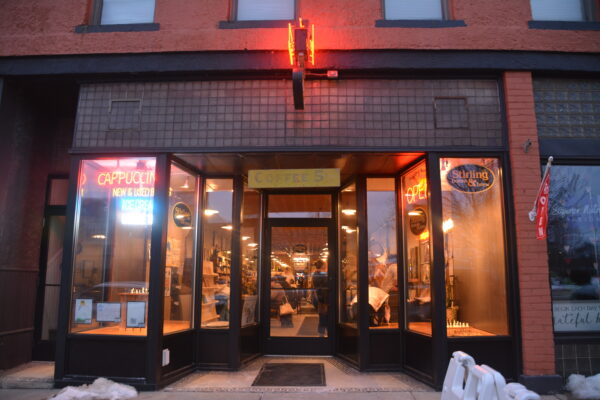 A storefront with floor-to-ceiling glass windows. Inside, people stand and talk or sit at tables, surrounded by warm light. Four signs hang at the front of the store. From left to right, neon signs read “Cappuccino New & Used Books,” “Coffee 5,” “Open” and “Stirling books & brew.”