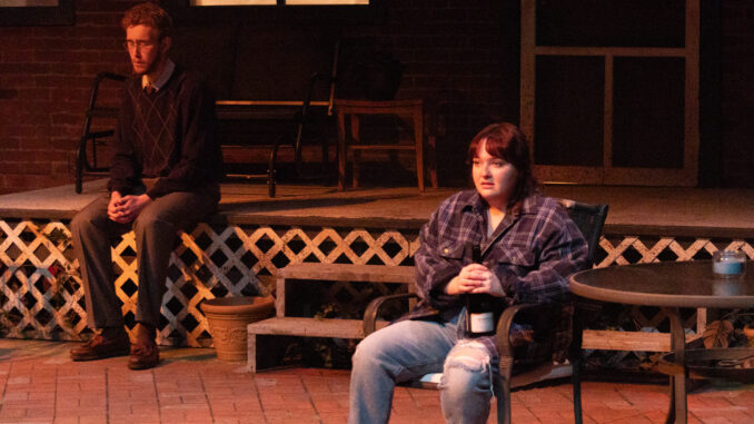 A man and a woman sit diagonally from each other, one on a raised platform and one in a metal chair, respectively. The woman is wearing a flannel with a champagne bottle in hand. The woman has a distraught expression on her face. In the back, the man sits on the steps of the house with his hands clasped together, with a solemn look on his face.